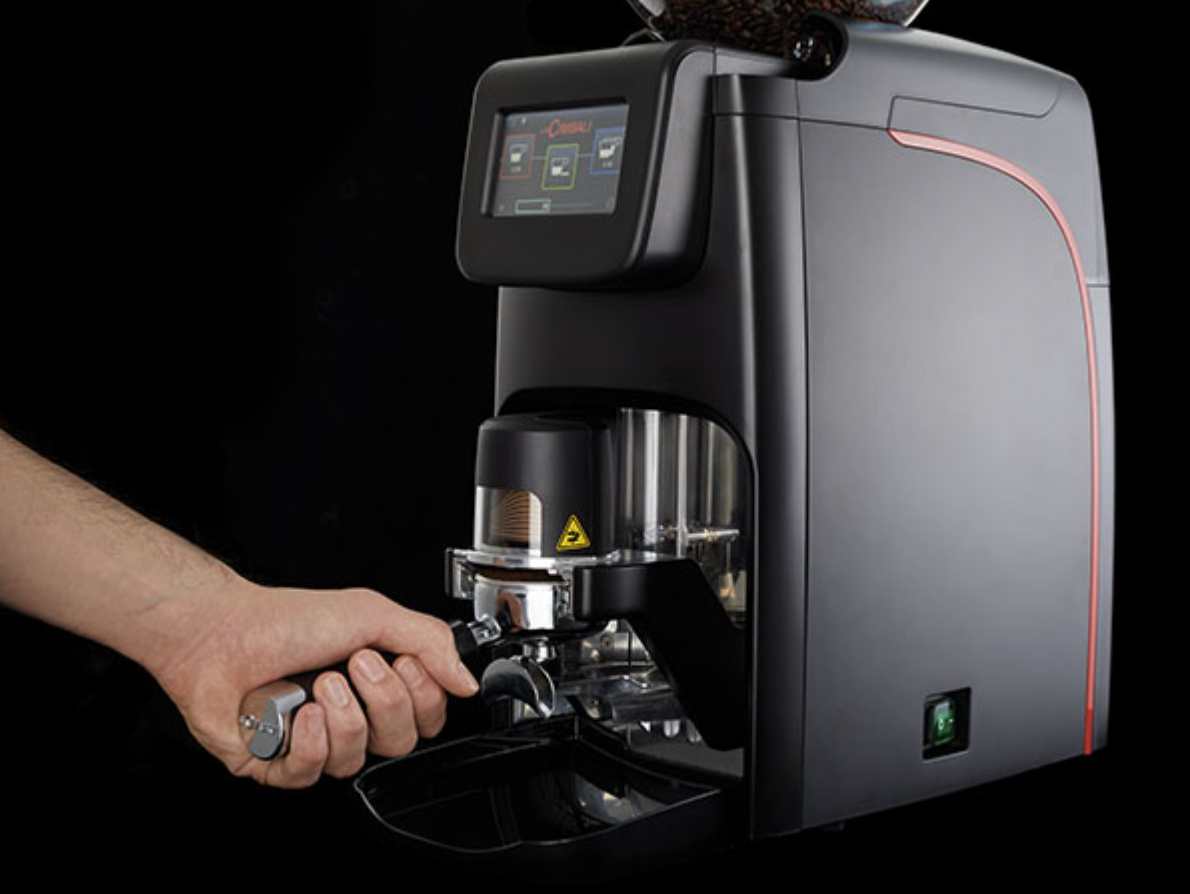 Barista using the black Cimbali Elective Auto Tamper grinder with portafilter during espresso preparation - Cimbali Elective Auto Tamper Grinder In Use – Black Edition - The Cimbali Elective Auto Tamper grinder in black delivers professional-grade performance with barista-friendly controls.
